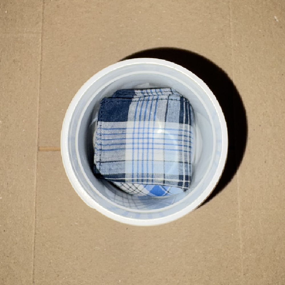 Top view of a folded blue and white plaid handkerchief inside a white capsule tub on a cardboard background.