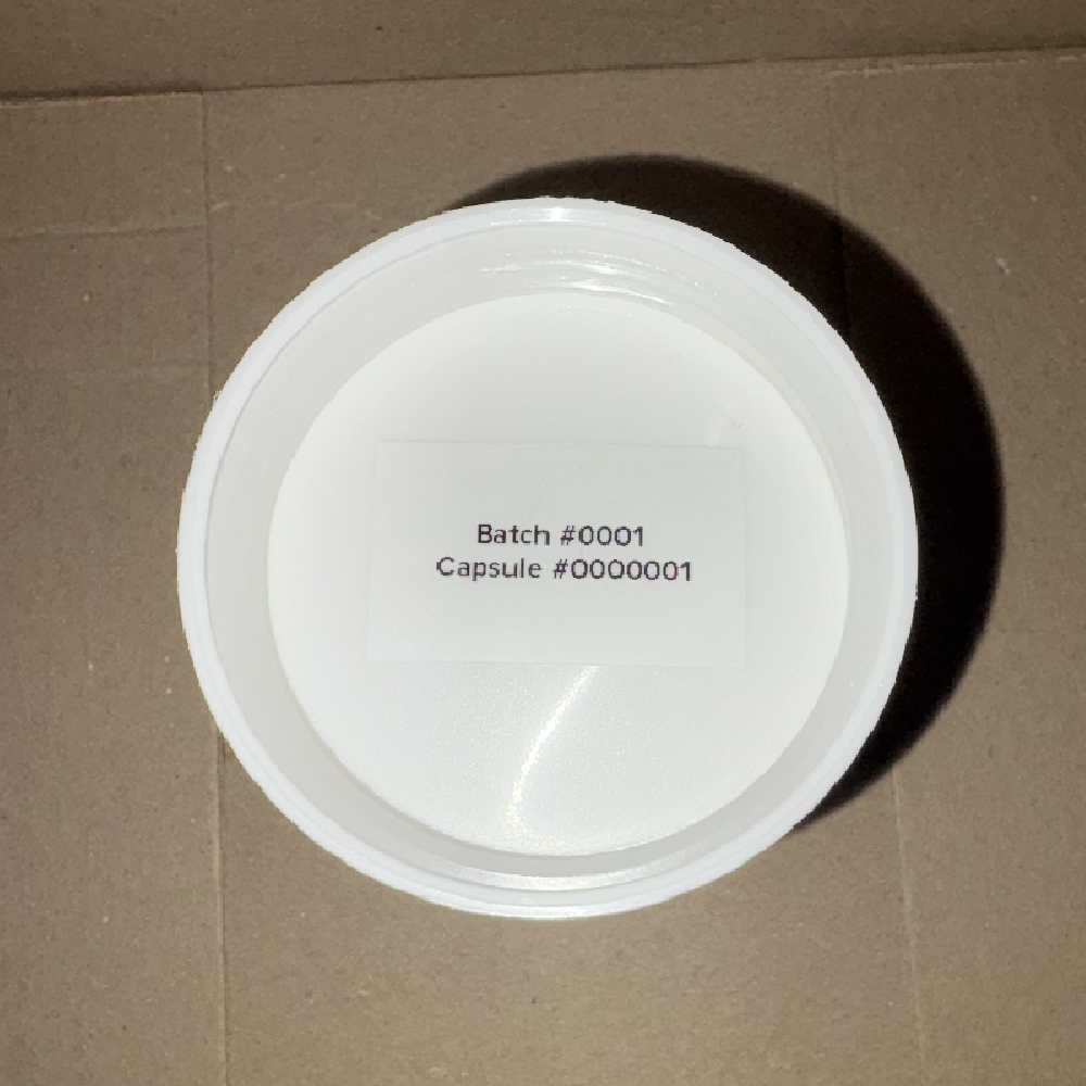 White capsule lid with printed label reading “Batch #0001, Capsule #0000001” on cardboard surface.